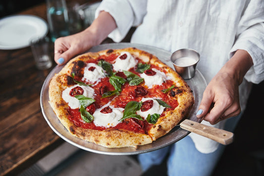 Pizzaiolo serve pizza gourmet con burrata, pomodorini e basilico su pala in pizzeria, focus sulla qualità degli ingredienti