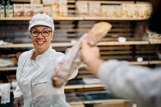 Panettiera sorridente consegna baguette a cliente in panificio moderno, scaffali con prodotti da forno sullo sfondo